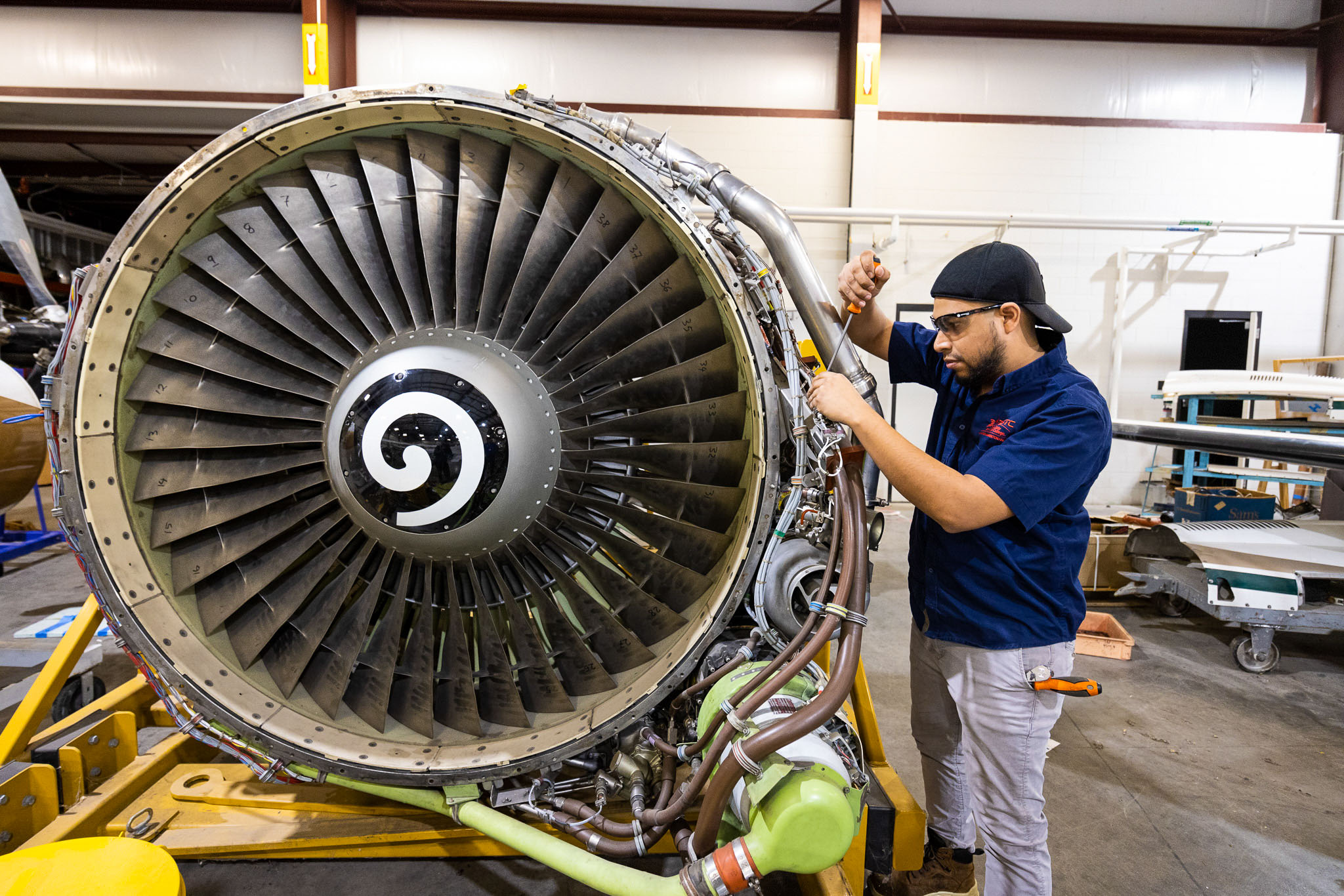 Technician in safety glasses uses a screwdriver to adjust wiring on a large exposed jet engine, with turbine blades prominently visible in an aviation workshop.